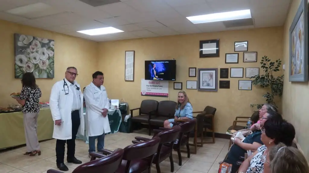 Clinic waiting room with medical staff, seated patients, TV, and framed certificates.